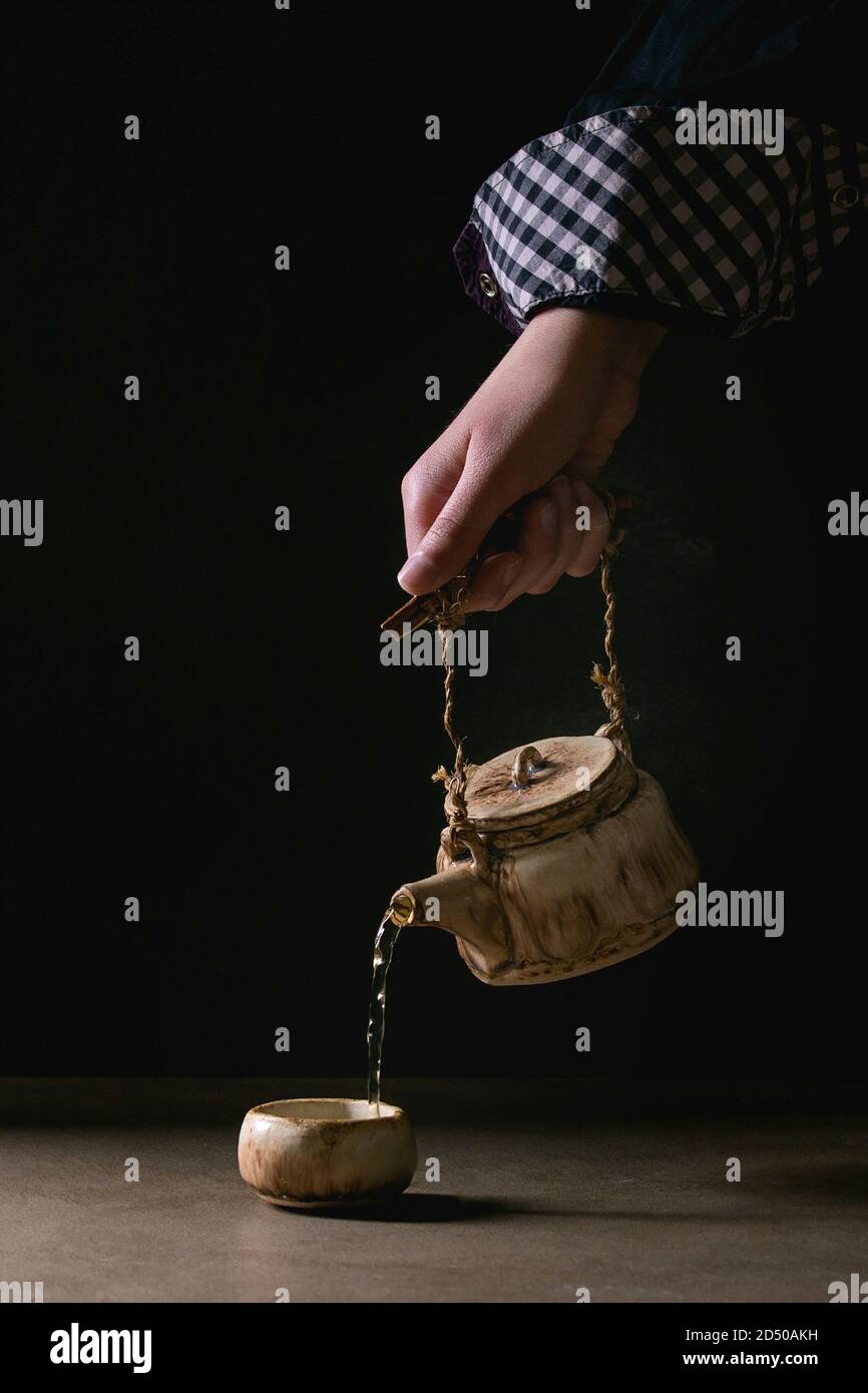 Jeune homme de de l'artisanat faits à la main chaude dans la théière de thé vert chinois traditionnel tasse en céramique argile debout sur le tableau sombre sur fond noir Banque D'Images