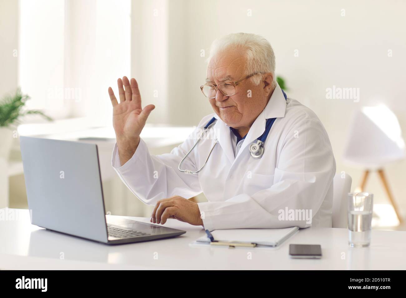 Un médecin expérimenté sympathique agite la main sur l'écran d'un ordinateur portable en commençant en ligne consultation avec le patient Banque D'Images
