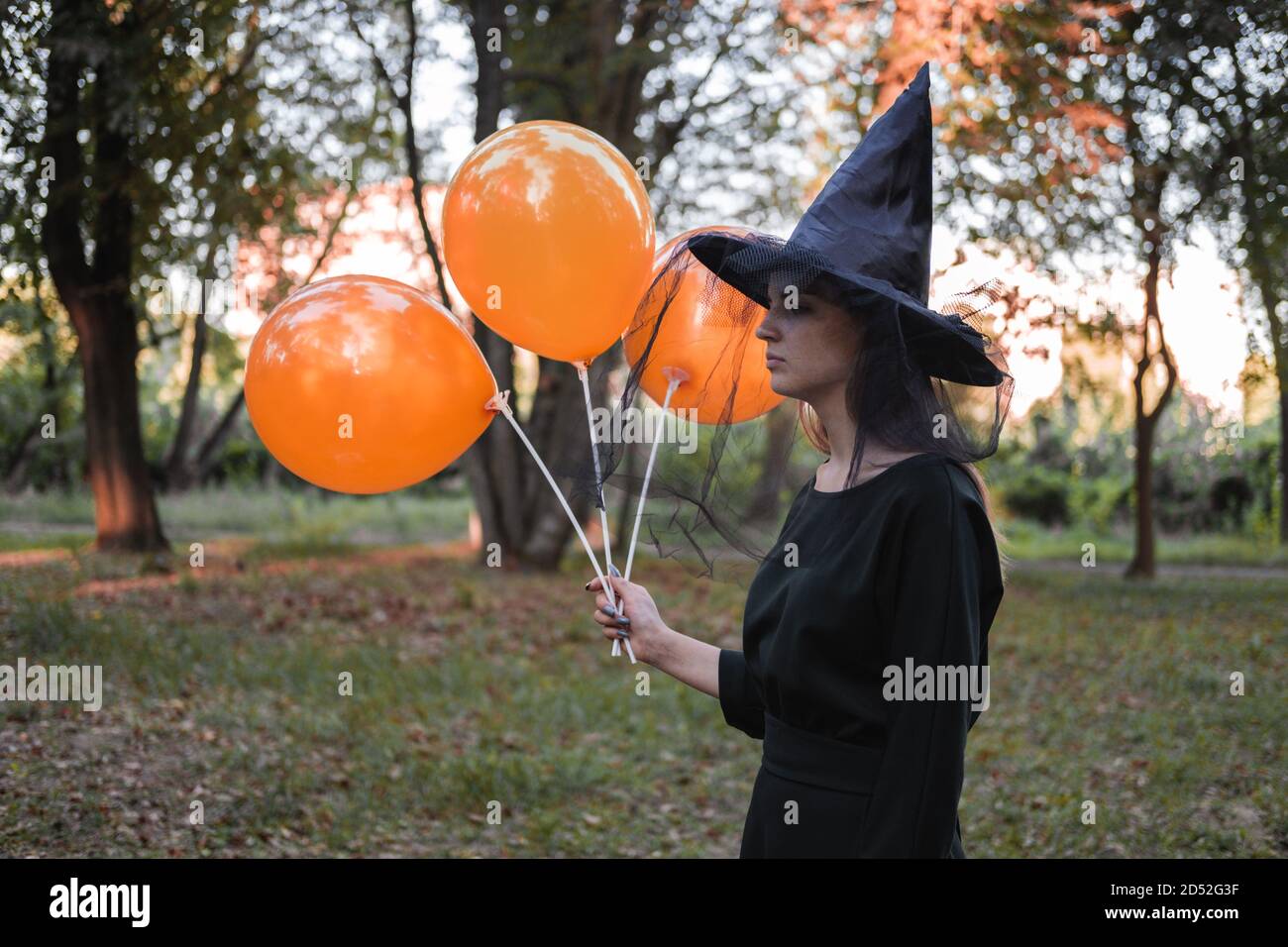 Jeune mignonne belle femme en robe sombre et chapeau de sorcière tient des ballons d'orange dans ses mains. Costume de fête d'Halloween. Forêt, parc avec arbres d'automne Banque D'Images