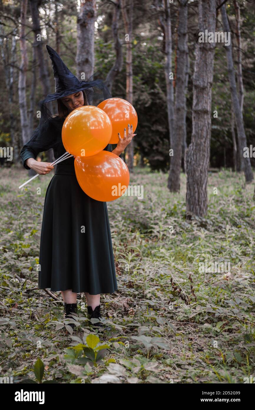 Jeune mignonne belle femme en robe sombre et chapeau de sorcière tient des ballons d'orange dans ses mains. Costume de fête d'Halloween. Forêt, parc avec arbres d'automne Banque D'Images