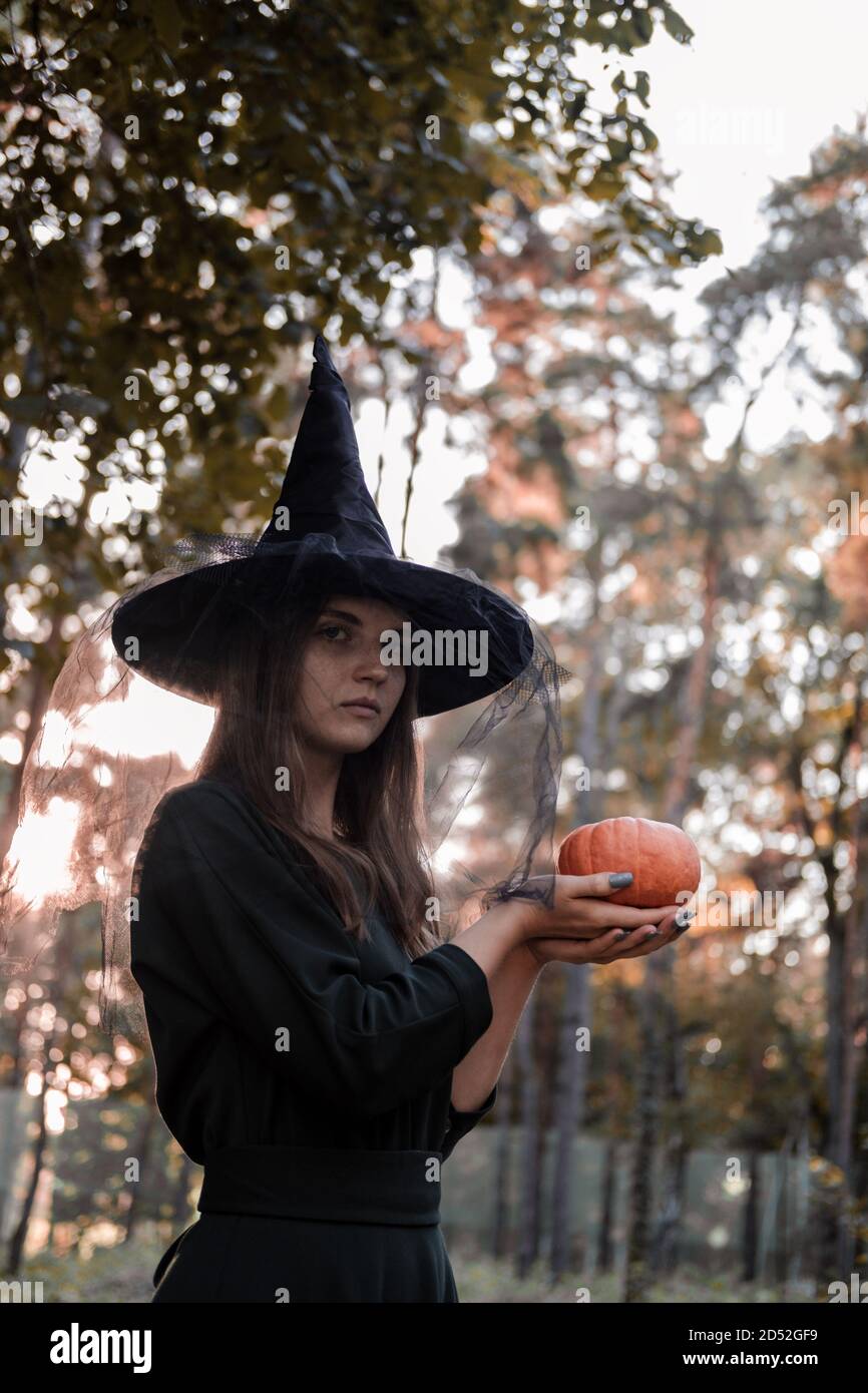 Une jeune femme en robe noire et chapeau de sorcière tient une citrouille dans ses mains. Costume de fête d'Halloween. Forêt, parc avec arbres d'automne Banque D'Images