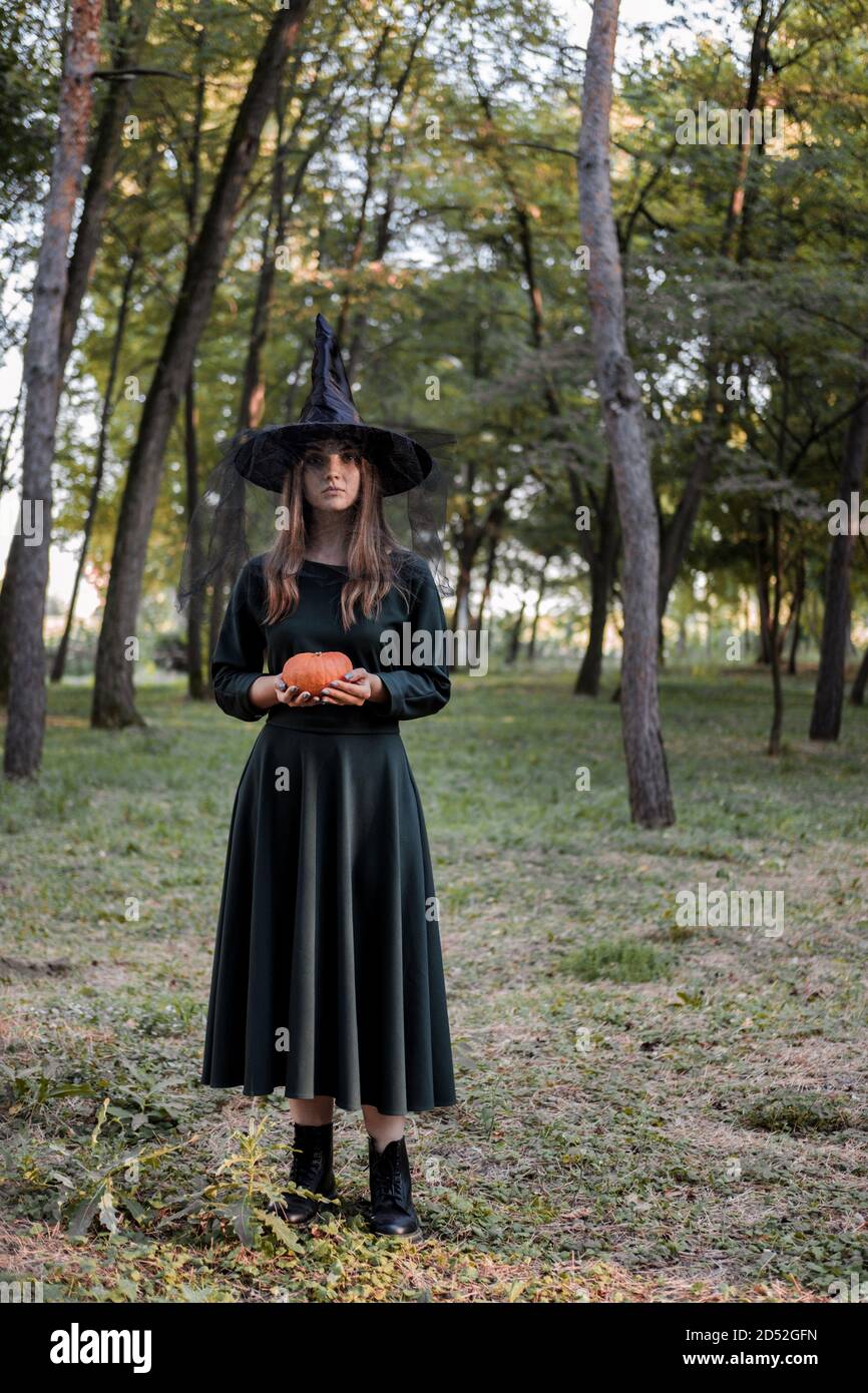 Une jeune femme en robe noire et chapeau de sorcière tient une citrouille dans ses mains. Costume de fête d'Halloween. Forêt, parc avec arbres d'automne Banque D'Images