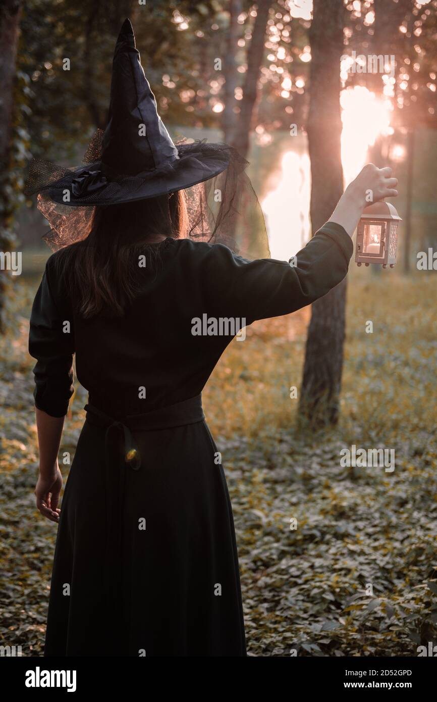 Une jeune femme en robe sombre et chapeau de sorcière tient une lanterne avec des bougies dans ses mains et illumine la forêt. Costume de fête d'Halloween. Garez-vous avec l'autom Banque D'Images