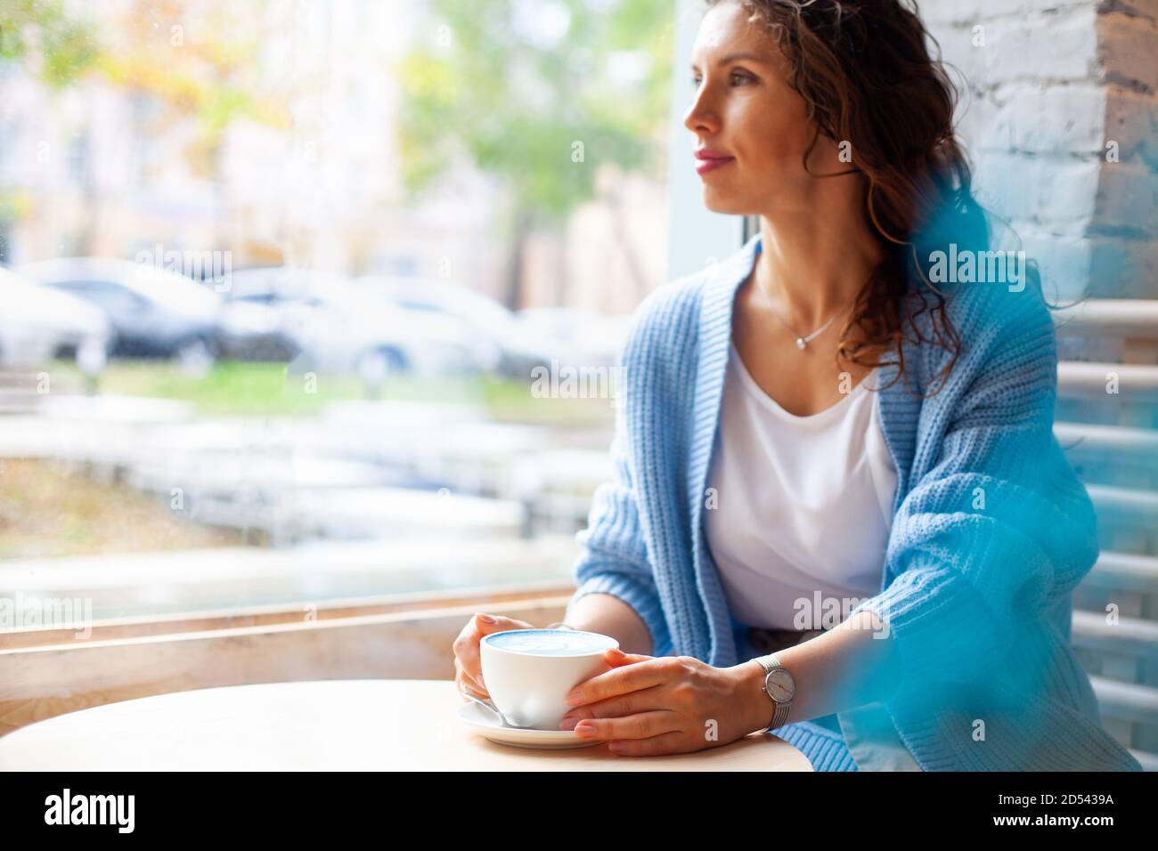 Femme heureuse non focalisée avec de longs cheveux ondulés dans un pull chaud contenant une tasse de lait bleu chaud Banque D'Images