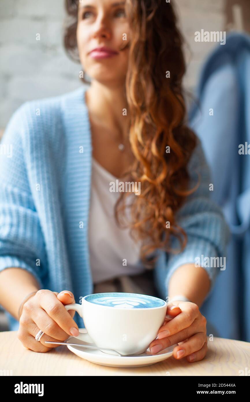Femme heureuse non focalisée avec de longs cheveux ondulés dans un pull chaud contenant une tasse de lait bleu chaud Banque D'Images