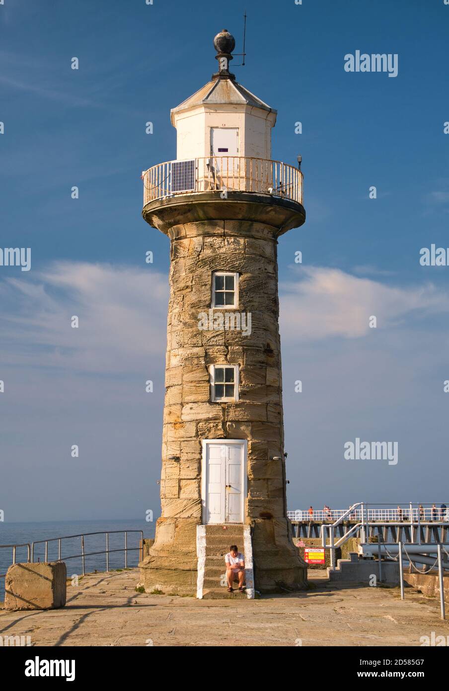 Un touriste regarde un téléphone tout en étant assis sur la pierre Marches au pied de l'historique grès Whitby West Pier Lighthouse dans un après-midi ensoleillé Banque D'Images