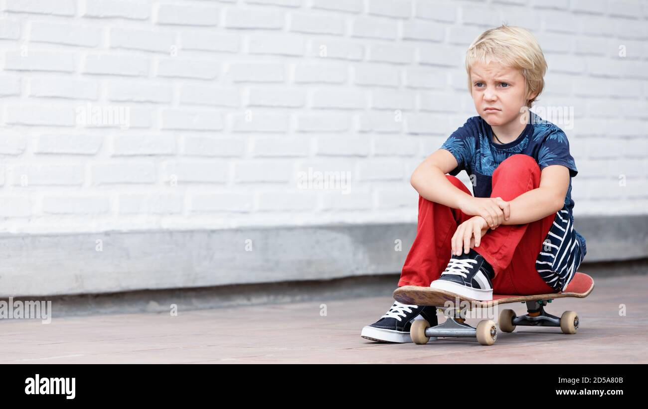Le patineur qui a l'air malheureux et contrarié s'assoit sur le skateboard avant que les enfants ne s'entraîne dans le parc de skate. Vie familiale active, activités de loisirs en plein air Banque D'Images