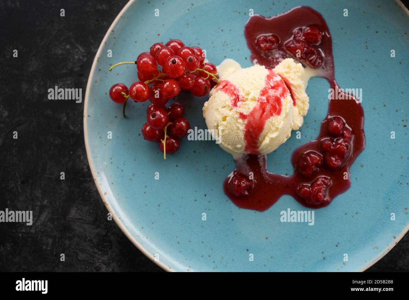 Cuillère à crème glacée à la vanille avec des raisins de Corinthe rouges et une sauce aux fruits sur une assiette bleue, fond sombre, espace de copie, vue en grand angle du dessus, mise au point sélectionnée Banque D'Images
