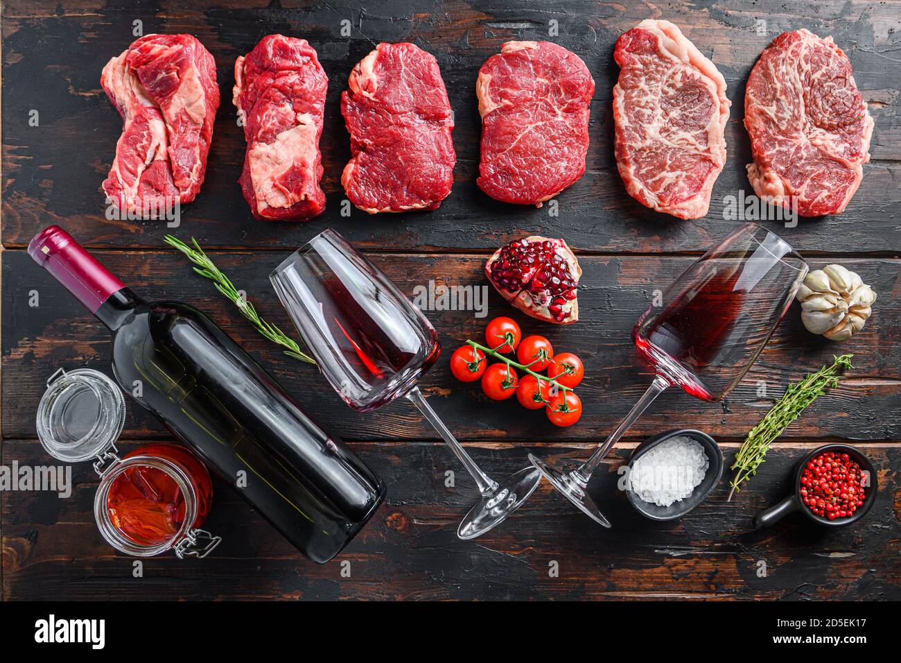 Ensemble de différents steaks de bœuf cru avec assaisonnements et vin rouge en bouteille et verre sur de vieilles planches sombres en bois vue de dessus. Banque D'Images