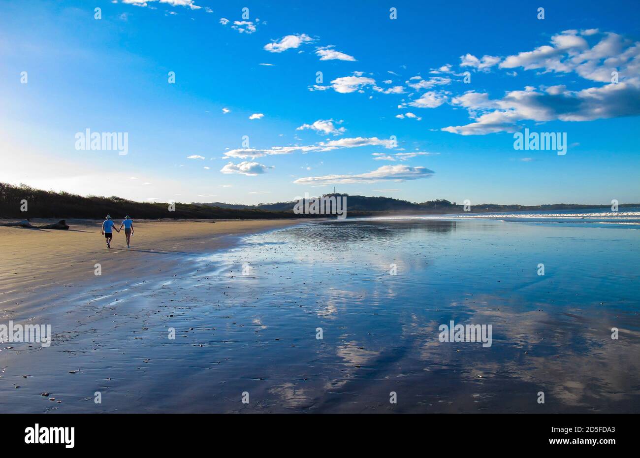 Deux personnes marchant le long de la plage de sable tropical avec un ciel bleu et des nuages blancs se reflétant dans l'eau avec des montagnes en arrière-plan Banque D'Images