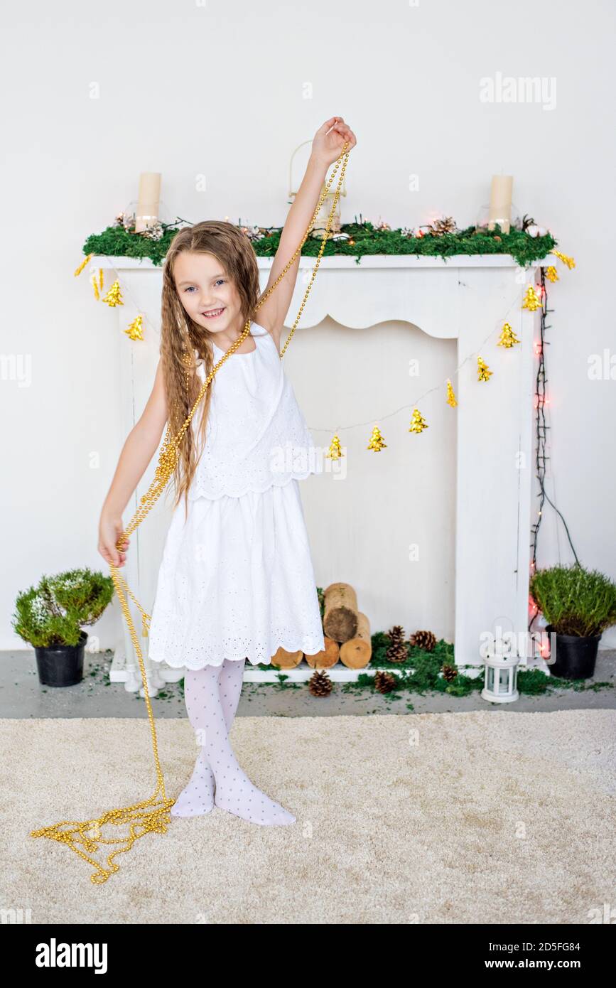 Portrait rapproché d'une petite fille blonde dans une robe blanche près d'un arbre de Noël avec une guirlande lumineuse. Vacances du nouvel an près de la cheminée à la maison. Banque D'Images