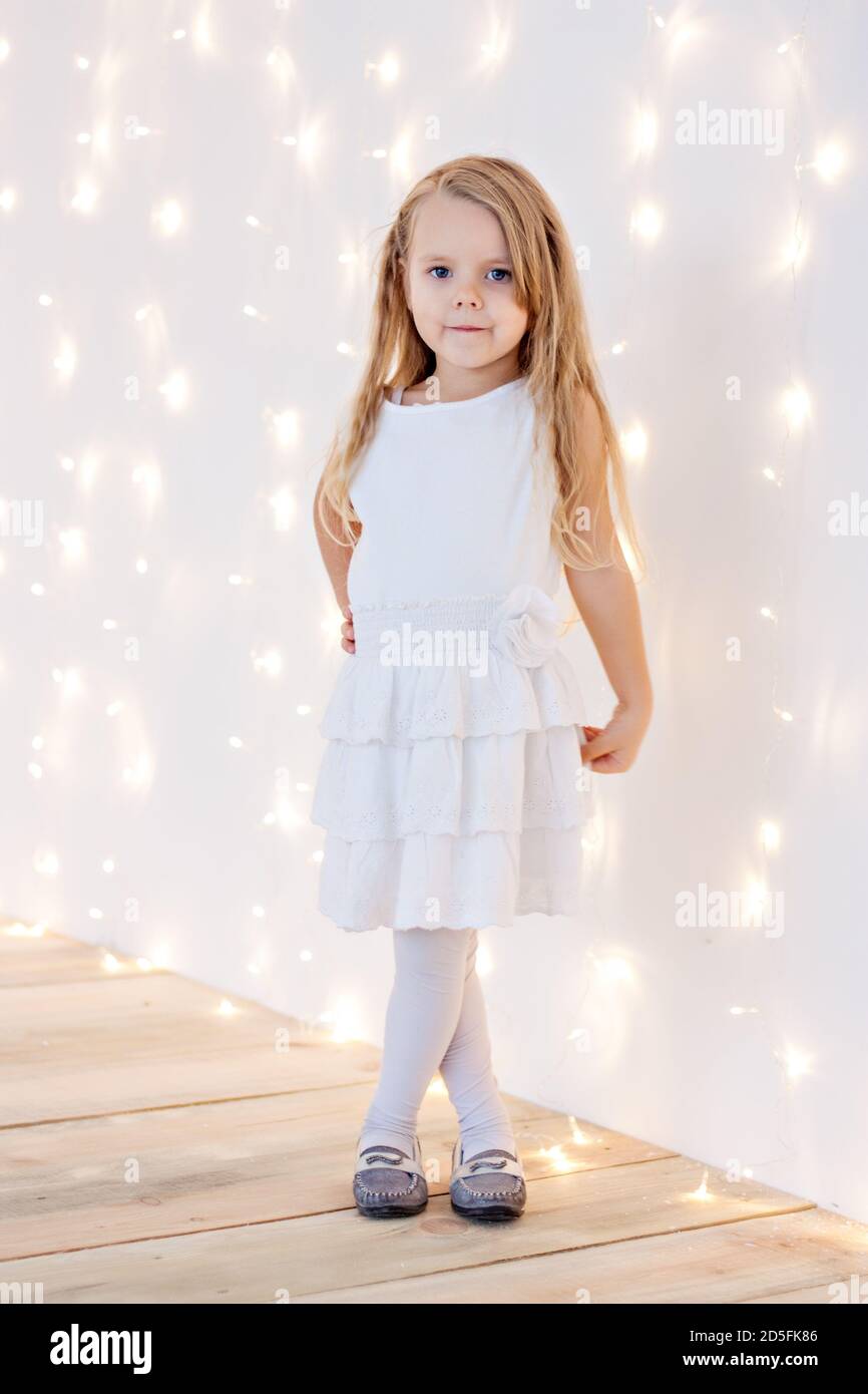 Portrait en gros plan d'une petite fille blonde dans une robe de fête blanche sur un fond de bokeh jaune provenant d'une guirlande de Noël. Carte du nouvel an. Bébé Banque D'Images