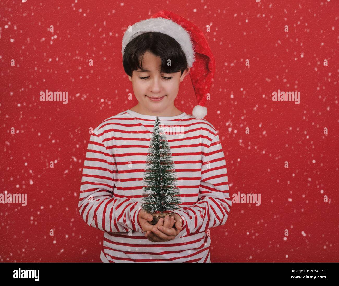 Joyeux Noël, un enfant portant un chapeau de Noël avec un petit arbre de noël entre ses mains sur fond rouge Banque D'Images