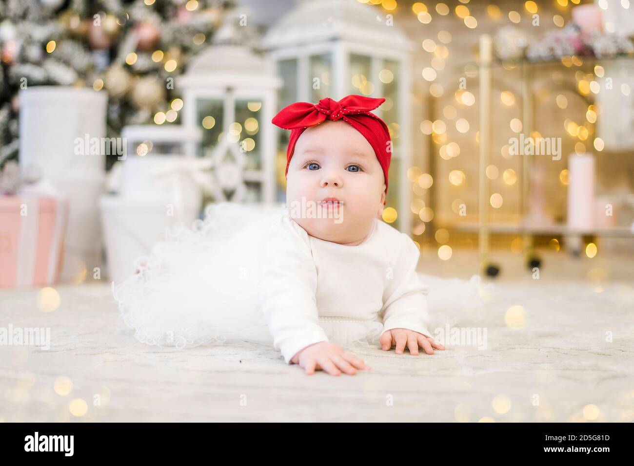 Portrait d'une petite fille dans une élégante robe blanche avec de la paille rouge sur sa tête et des chaussons sur le fond d'un luxueux arbre de Noël, firemplacer Banque D'Images