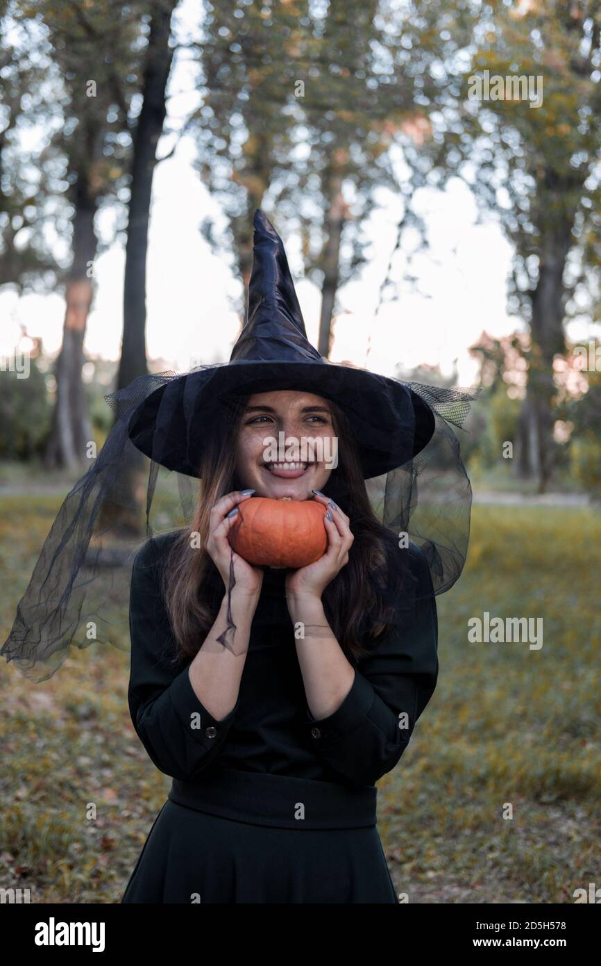 Une jeune femme en robe noire et chapeau de sorcière tient une citrouille dans ses mains. Costume de fête d'Halloween. Forêt, parc avec arbres d'automne Banque D'Images