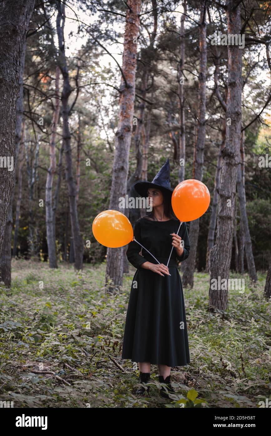Jeune mignonne belle femme en robe sombre et chapeau de sorcière tient des ballons d'orange dans ses mains. Costume de fête d'Halloween. Forêt, parc avec arbres d'automne Banque D'Images