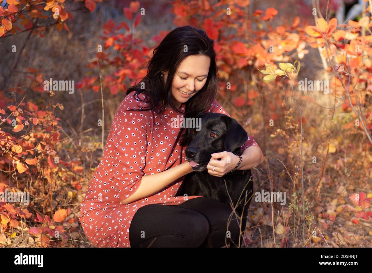 La jeune femme asiatique en robe rouge joue avec le Labrador noir en forêt d'automne Banque D'Images