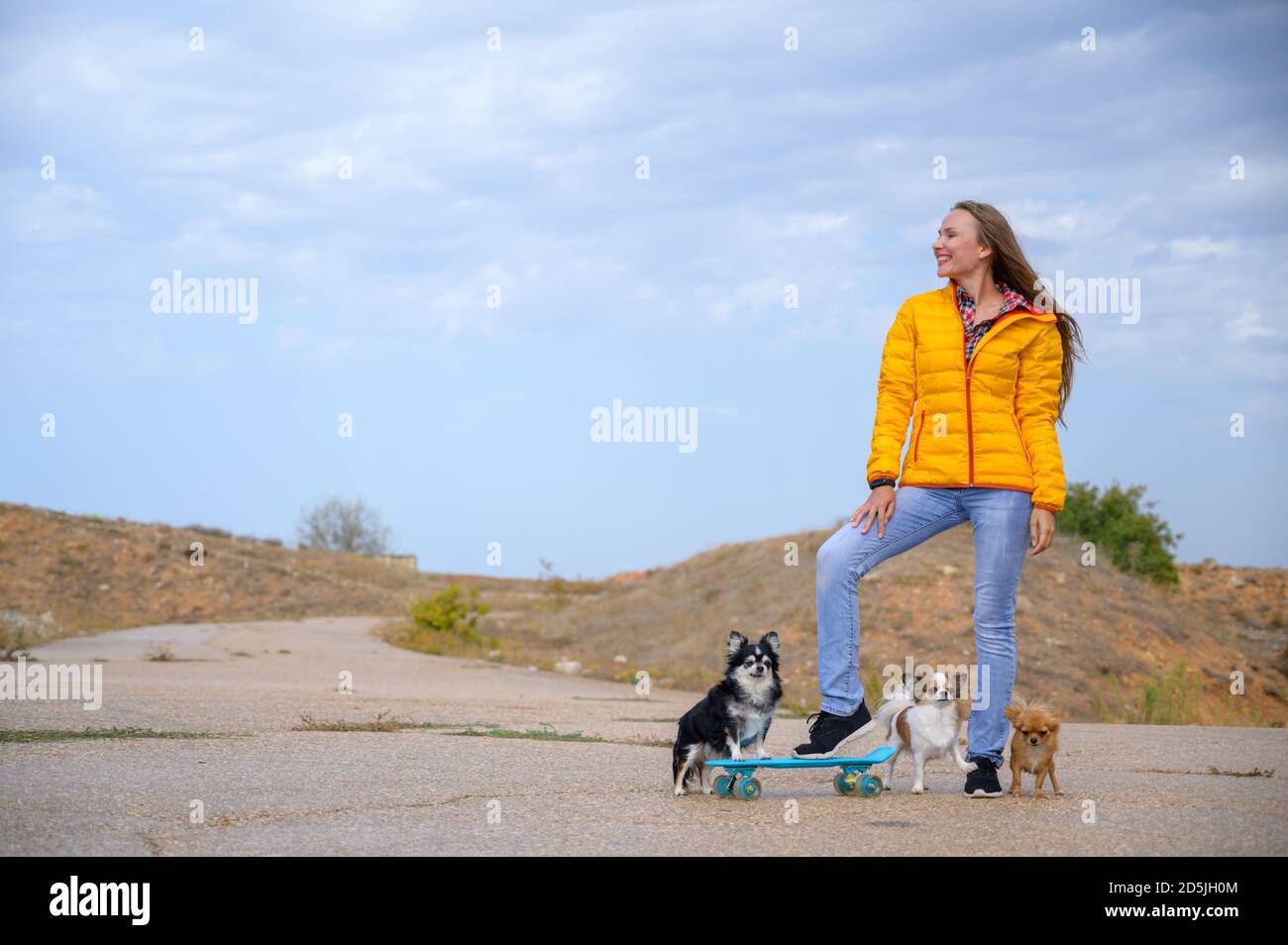active sport fille en veste jaune automne et jeans bleu debout avec un skateboard et trois chiens chihuahua sur le ciel nuageux fond ciel Banque D'Images