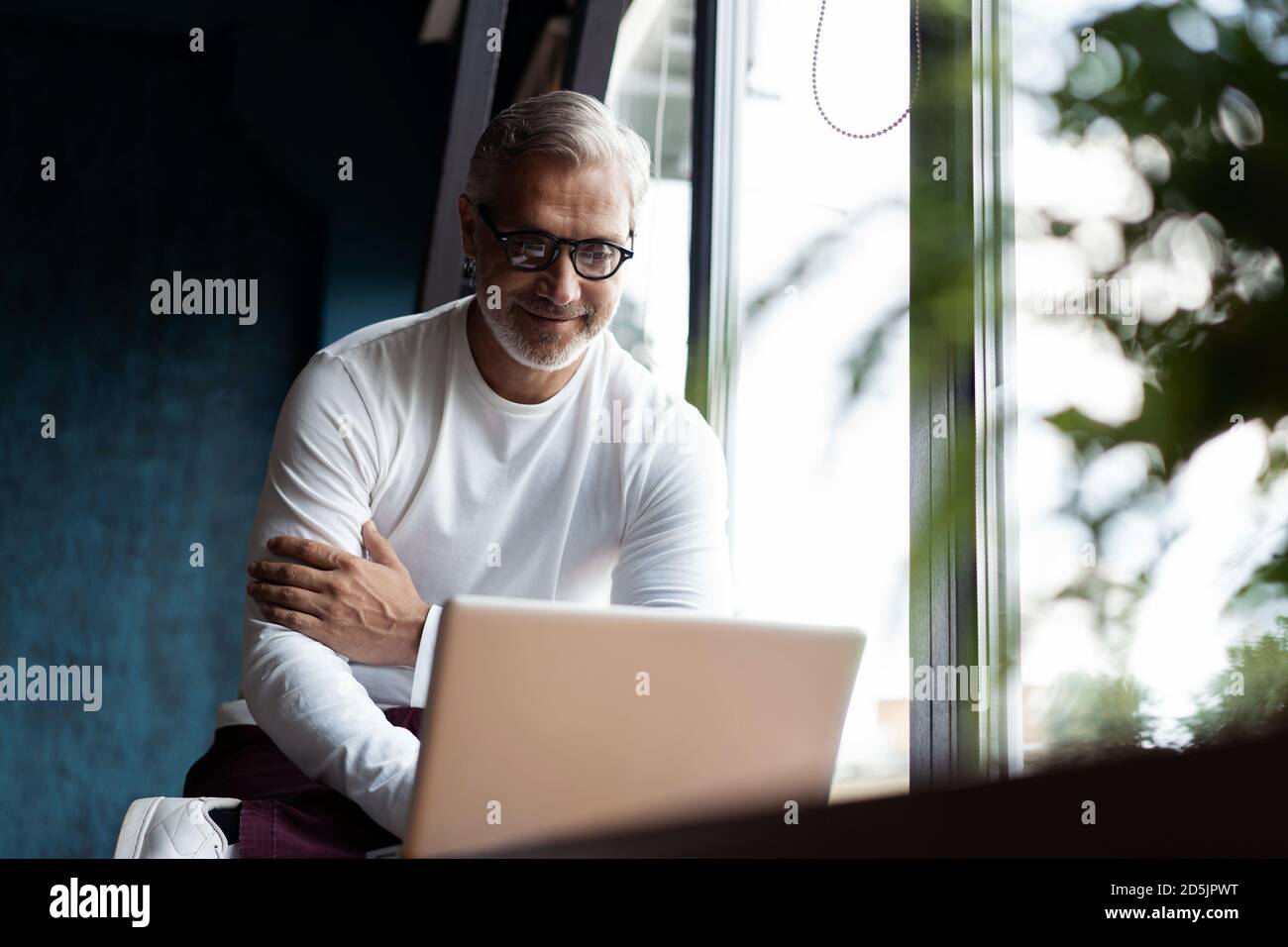 Décontracté gris-cheveux homme mature dans des lunettes de vue travaillant sur ordinateur portable pendant assis dans un bureau ou un café près de la fenêtre Banque D'Images