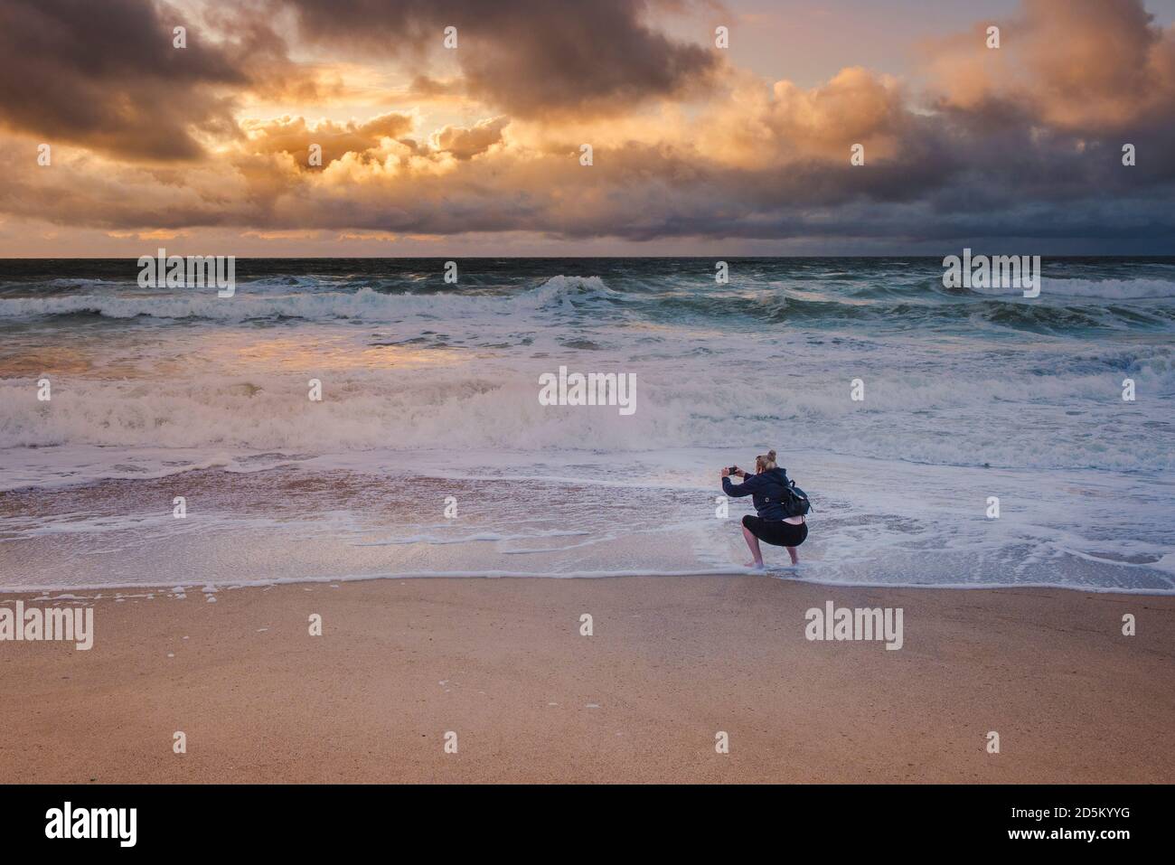 Un holidaymaker utilisant son téléphone portable pour photographier un coucher de soleil spectaculaire sur la plage de Fistral à Newquay en Cornouailles. Banque D'Images