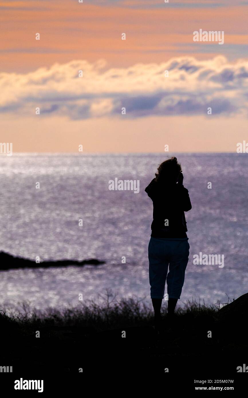 Une femme se tenant sur Pentire point East, regardant la mer et silhouetée par un coucher de soleil coloré à Newqiau dans les Cornouailles. Banque D'Images