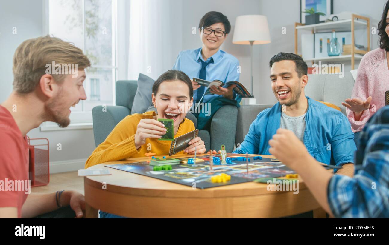 Groupe diversifié de gars et de filles jouant dans un jeu de plateau stratégique avec cartes et Dice. L'homme lance Dice de façon impressionnante. Salon confortable en journée Banque D'Images