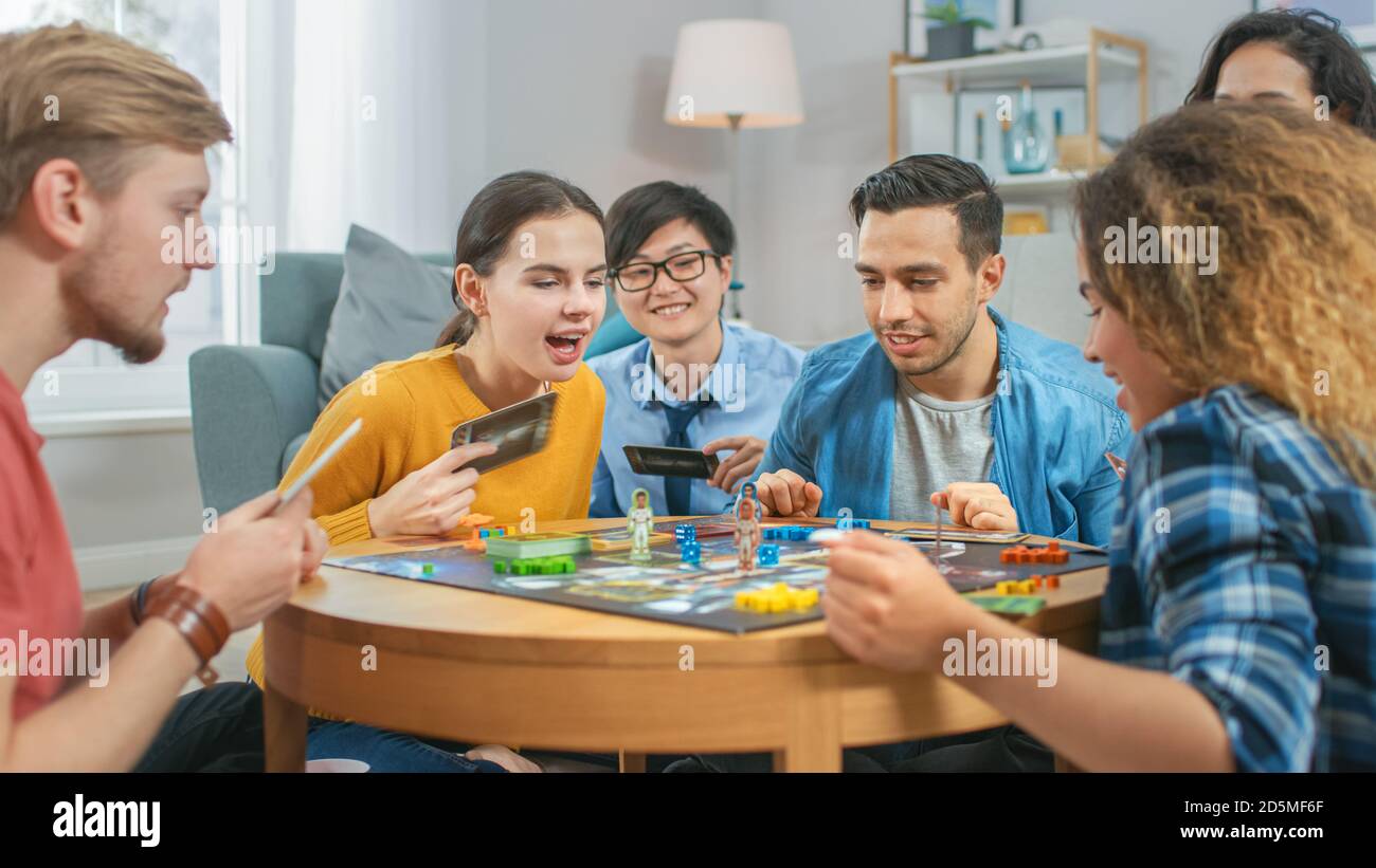 Groupe diversifié de gars et de filles jouant dans un jeu de plateau stratégique avec cartes et Dice. Salon confortable en journée Banque D'Images