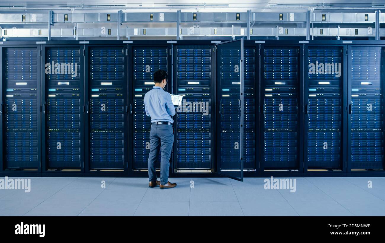 Dans le centre de données moderne : l'ingénieur INFORMATIQUE se trouve à côté des armoires rack de serveur ouvertes, effectue la procédure de maintenance et de diagnostic sans fil avec un ordinateur portable. Banque D'Images