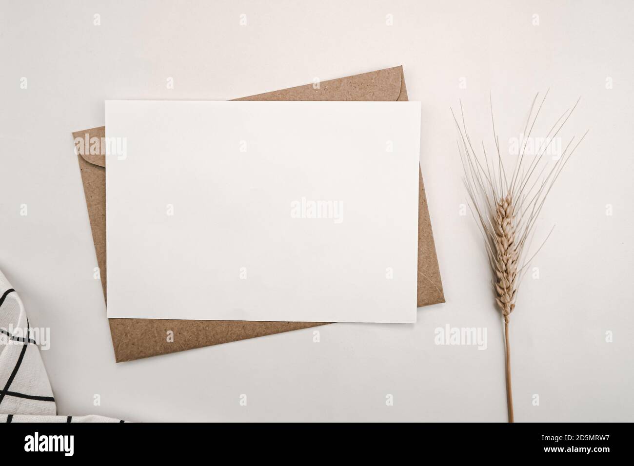 Papier blanc vierge sur enveloppe en papier marron avec fleur d'orge sèche et tissu blanc avec motif de grille noir. Maquette de carte de vœux vierge horizontale. T Banque D'Images