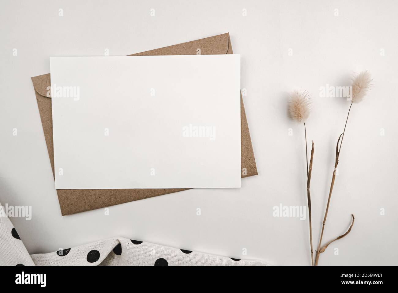 Papier blanc vierge sur enveloppe en papier marron avec fleur sèche de queue de lapin et tissu blanc avec points noirs. Maquette de carte de vœux vierge horizontale. Haut Banque D'Images
