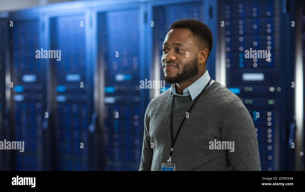 Technicien informatique de centre de données noir au milieu d'un couloir de rack de serveur. Il sourit. Banque D'Images