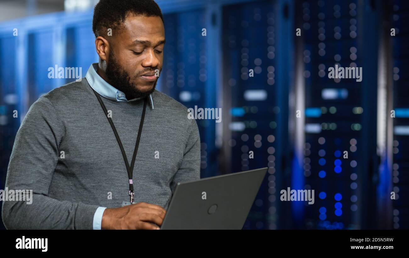 Technicien informatique de centre de données noir dans le couloir du rack de serveurs avec un ordinateur portable. Il inspecte visuellement les armoires de serveur de travail. Banque D'Images