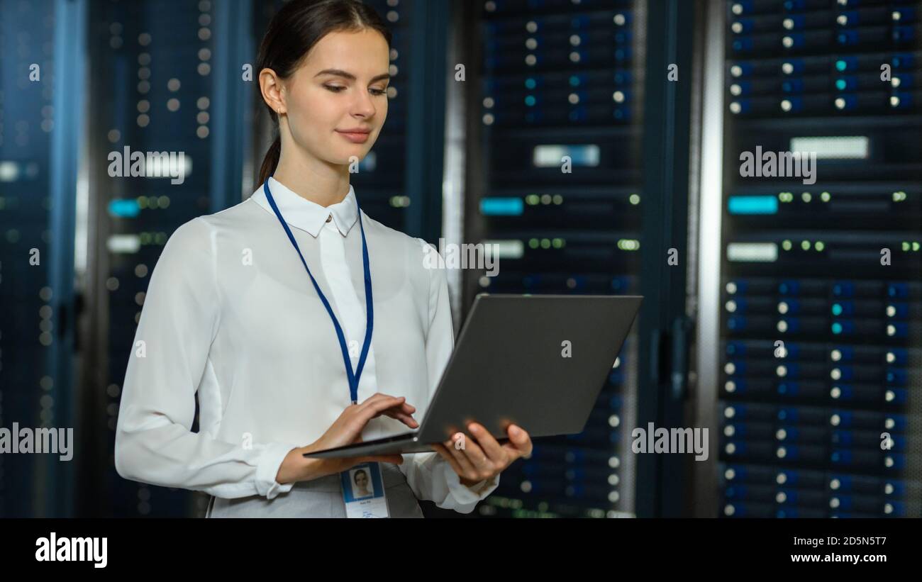 Magnifique technicien INFORMATIQUE féminin de centre de données situé près du couloir du rack de serveurs avec un ordinateur portable. Elle utilise un ordinateur portable et travaille avec des serveurs. Banque D'Images