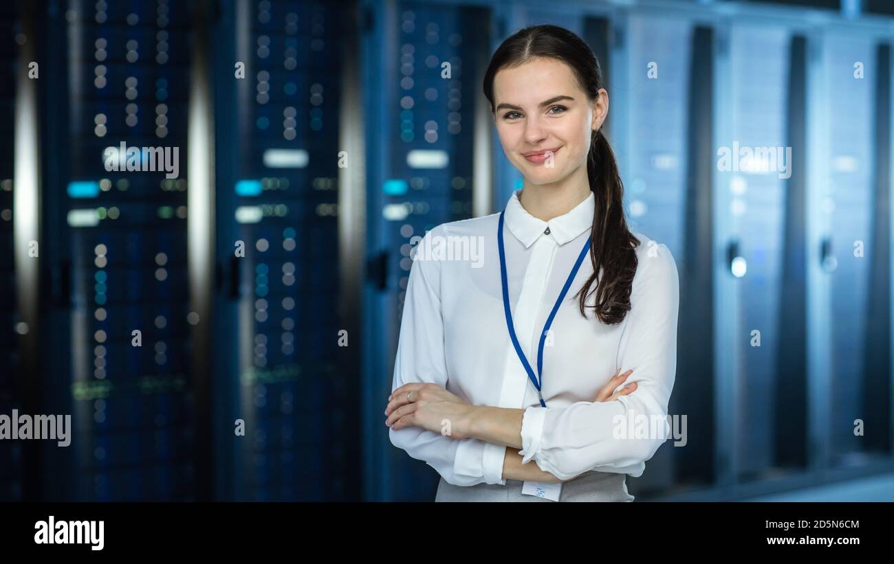 Une femme spécialiste INFORMATIQUE se trouve à la caméra du datacenter, à côté des racks de serveurs, et regarde la caméra. Ses bras ont traversé et elle Smiles à la Banque D'Images
