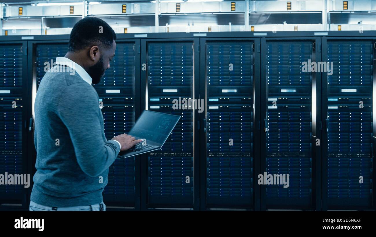 Technicien informatique de centre de données noir traversant le couloir du rack de serveurs avec un ordinateur portable. Il inspecte visuellement les armoires de serveur de travail. Banque D'Images