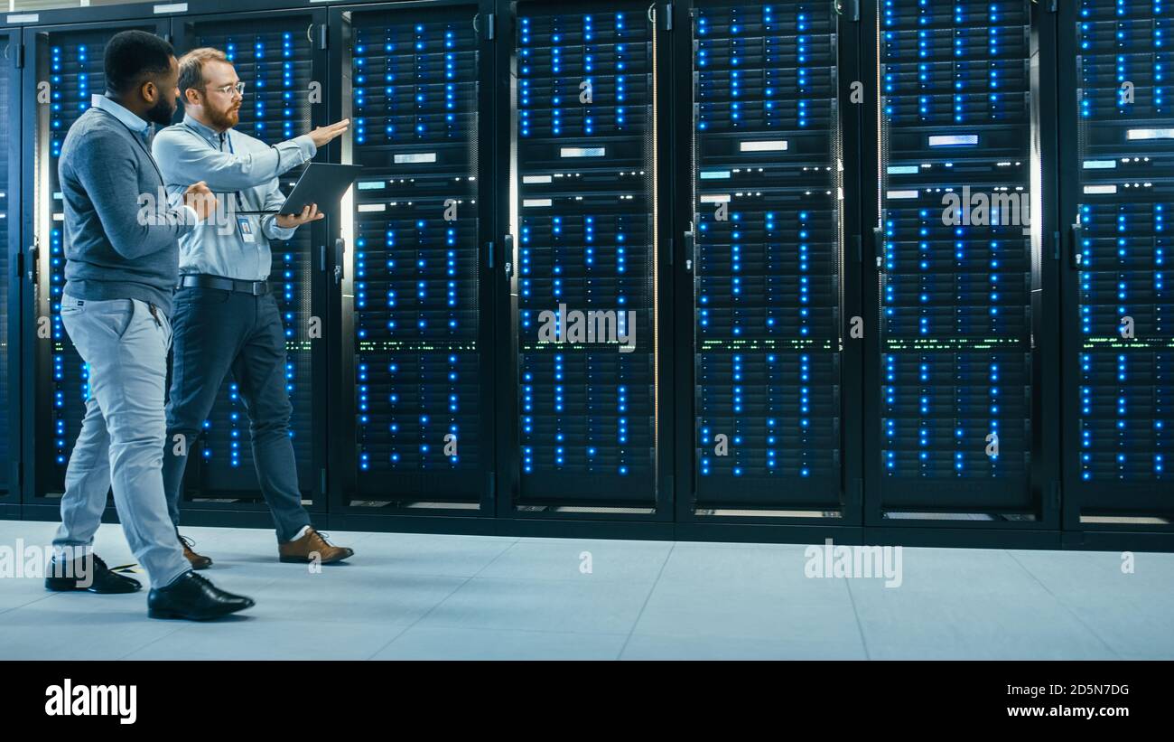 Un technicien INFORMATIQUE équipé d'un ordinateur portable et un collègue d'ingénieur noir parlent dans un centre de données lorsqu'il se trouve à côté des racks de serveurs. En cours d'exécution Banque D'Images