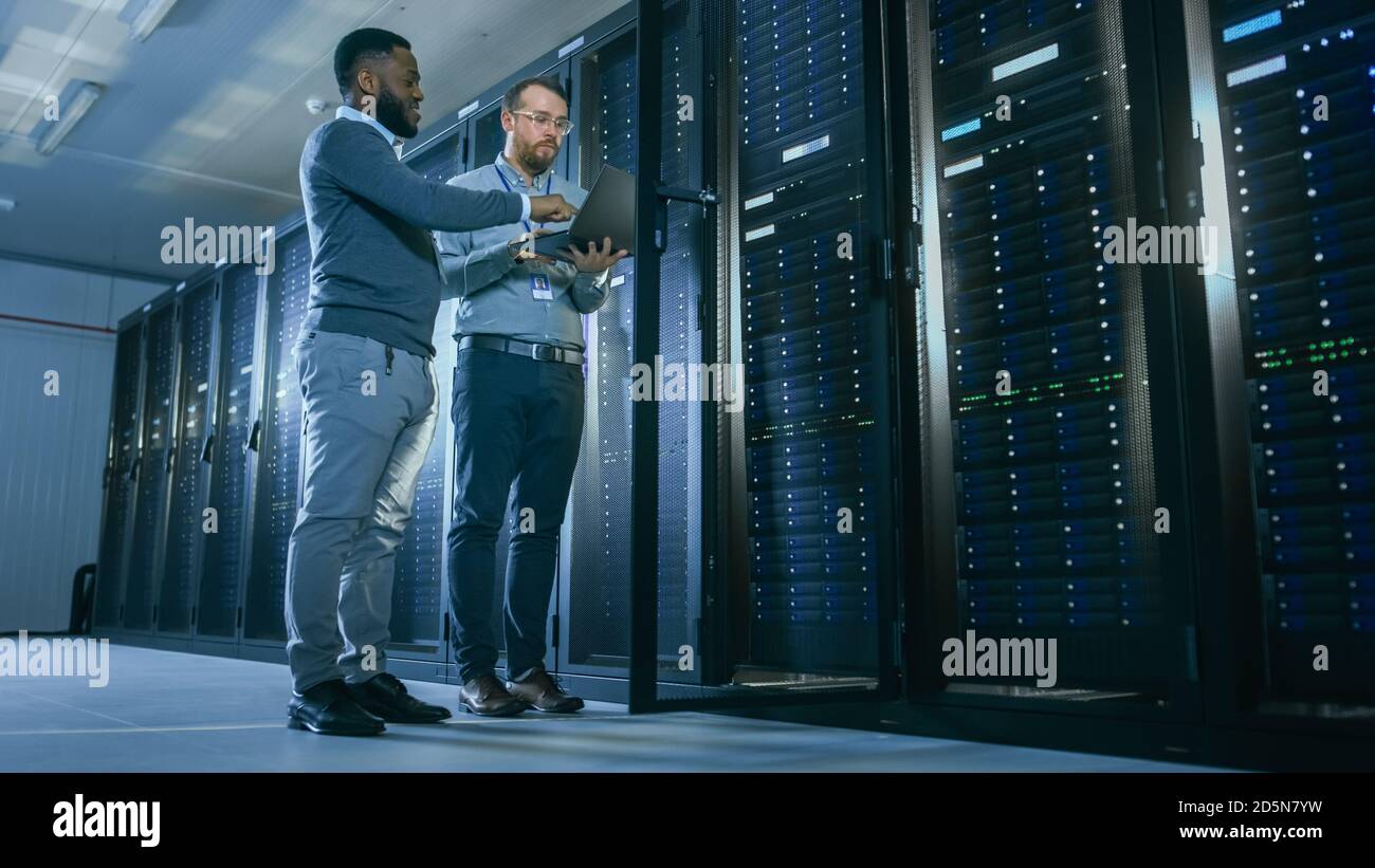 Technicien INFORMATIQUE barbu dans des lunettes avec un ordinateur portable et Un collègue ingénieur noir parle dans un centre de données pendant Travail à côté du serveur Banque D'Images