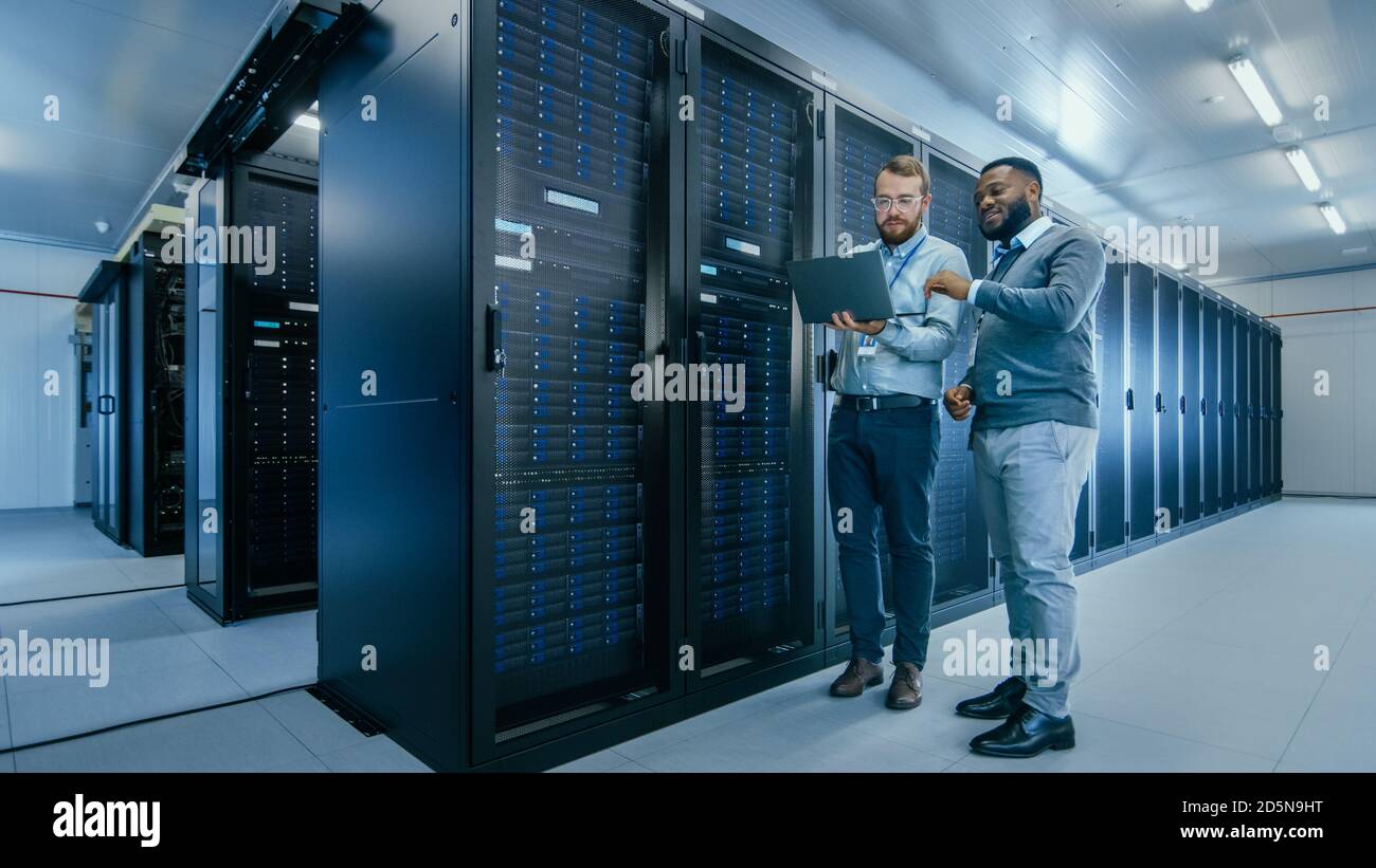Technicien INFORMATIQUE barbu dans des lunettes avec un ordinateur portable et Un collègue ingénieur noir parle dans un centre de données pendant Travail à côté du serveur Banque D'Images
