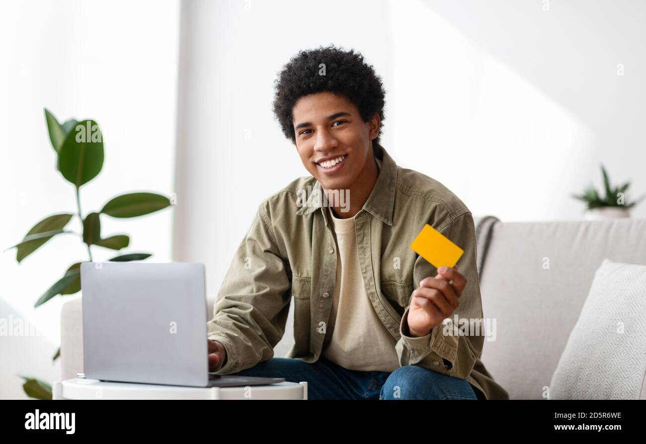 Concept Cybershopping. Homme adolescent noir souriant avec ordinateur portable et carte de crédit commandant des marchandises en ligne de la maison Banque D'Images