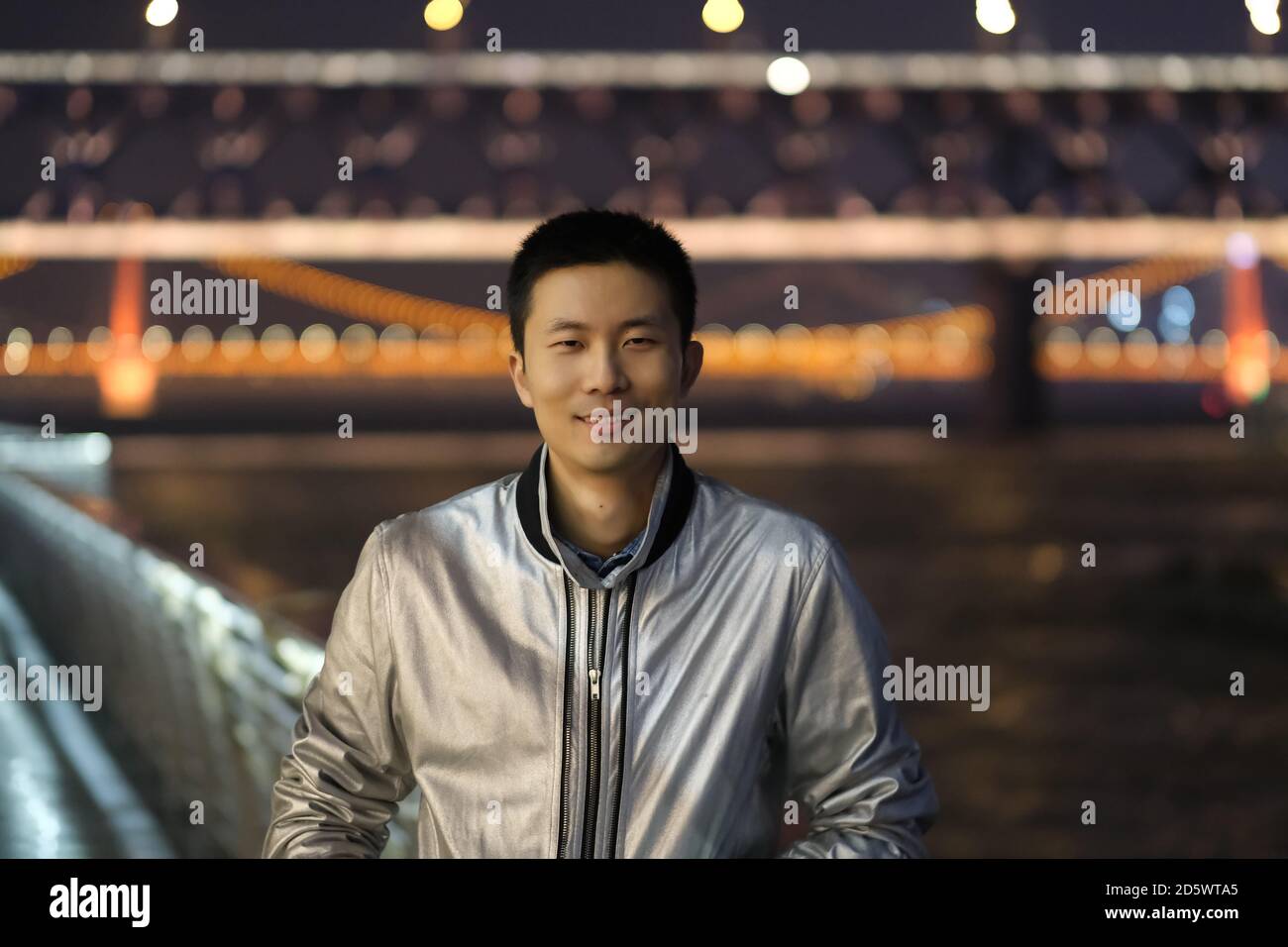 Prise de vue moyenne d'un jeune homme asiatique regardant l'appareil photo la nuit. Sourire heureux. Le pont du fleuve Yangtze de Wuhan a été défoqué comme arrière-plan Banque D'Images