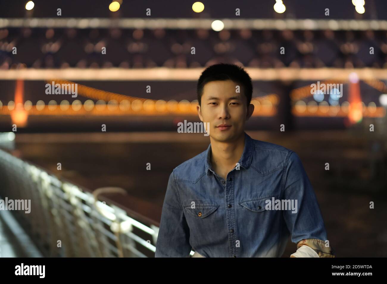 Prise de vue moyenne d'un jeune homme asiatique regardant l'appareil photo la nuit. Le pont du fleuve Yangtze de Wuhan a été défoqué comme arrière-plan Banque D'Images