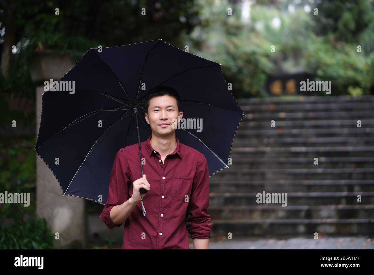 Prise de vue moyenne d'un jeune homme asiatique regardant la caméra, sous un parapluie et un sourire. Dans le parc naturel Banque D'Images