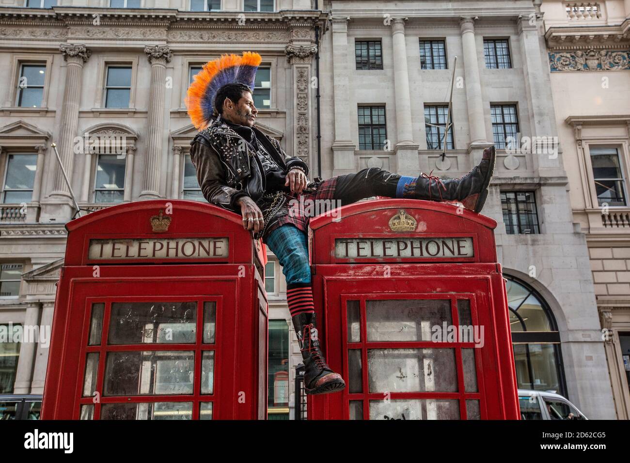 Punk rocker avec des cheveux mohican se détend sur le dessus des boîtes téléphoniques rouges dans le centre de Londres, Angleterre, Royaume-Uni Banque D'Images