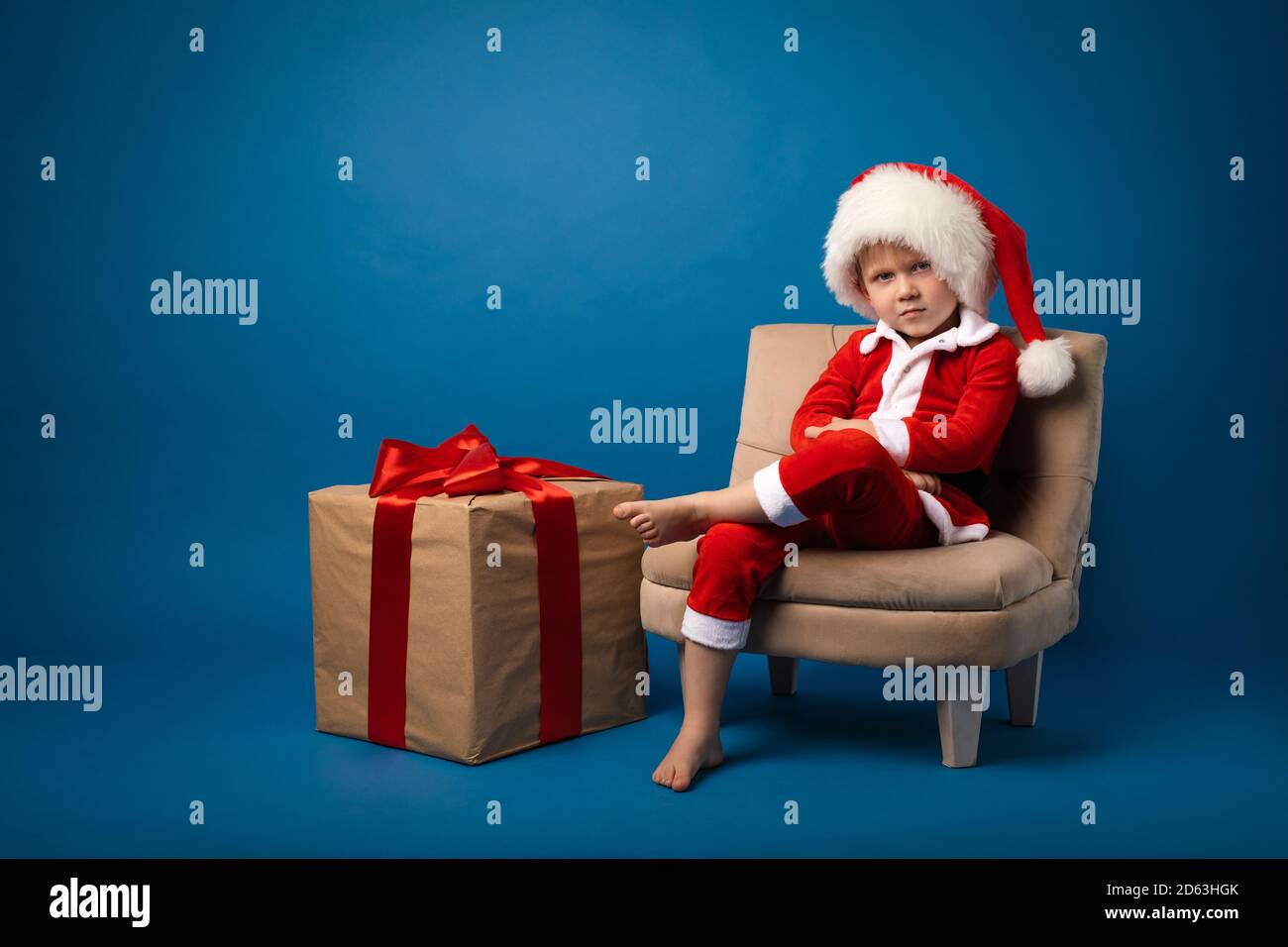 Un enfant garçon dans un mauvais costume de santa enfant avec un chapeau de nouvel an . Une immense boîte cadeau avec un arrière-plan bleu ruban rouge. Holiday concept et félicitations, stu Banque D'Images
