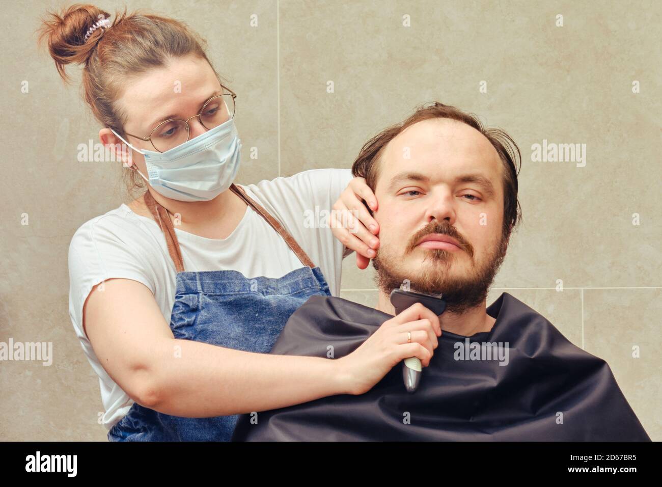 Le coiffeur dans un masque médical avec une tondeuse coupe la barbe d'un  client dans la salle de bains à la maison. Concept de coiffeurs et de  barbershops fermés dans l'isolement f