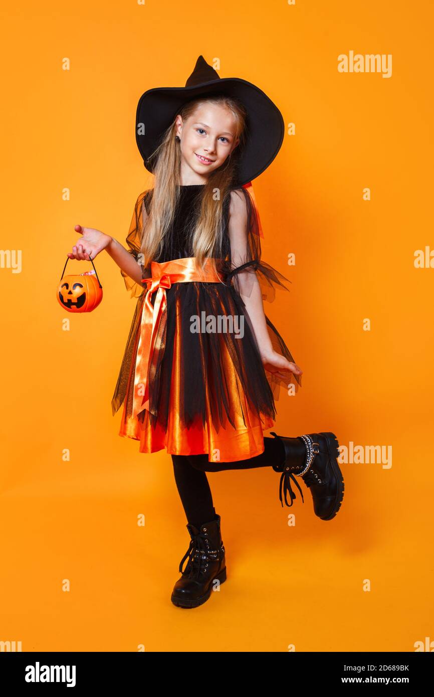 Jolie petite fille heureuse vêtue comme sorcière avec seau de bonbons sur fond orange Banque D'Images