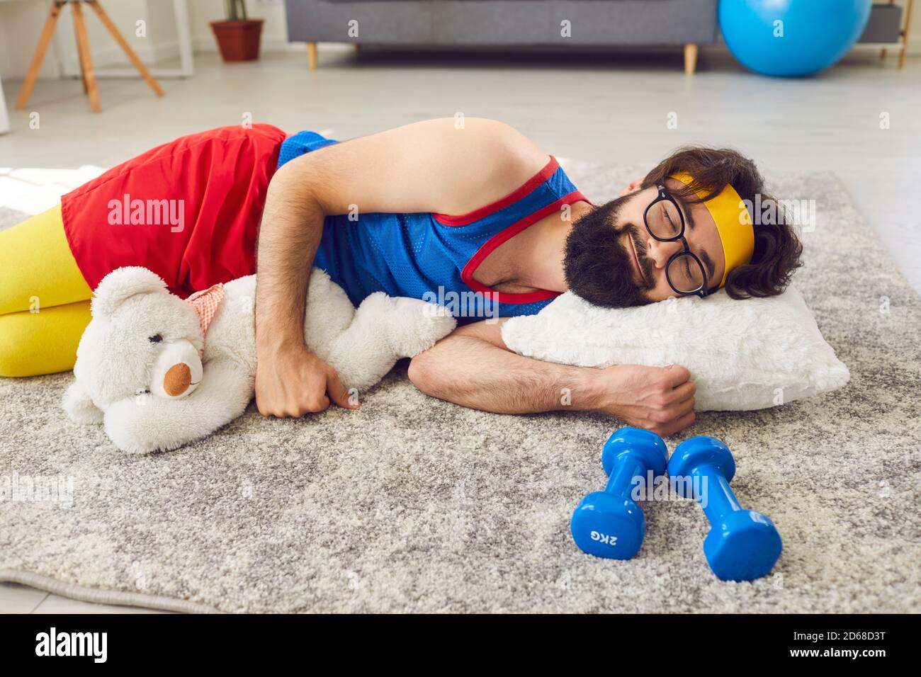 Athlète paresseux dormant paisiblement sur le sol, embrassant un ours en peluche, avec des haltères à côté Banque D'Images
