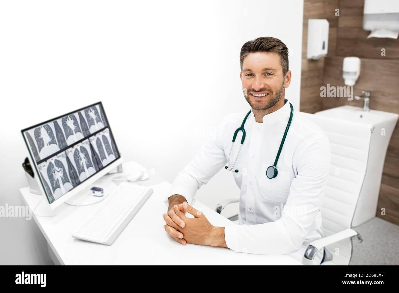 Portrait d'un médecin sur son lieu de travail dans une clinique moderne. Thérapeute avec un stéthoscope assis dans un bureau médical. Une radiographie des poumons se trouve sur son ordinateur Banque D'Images