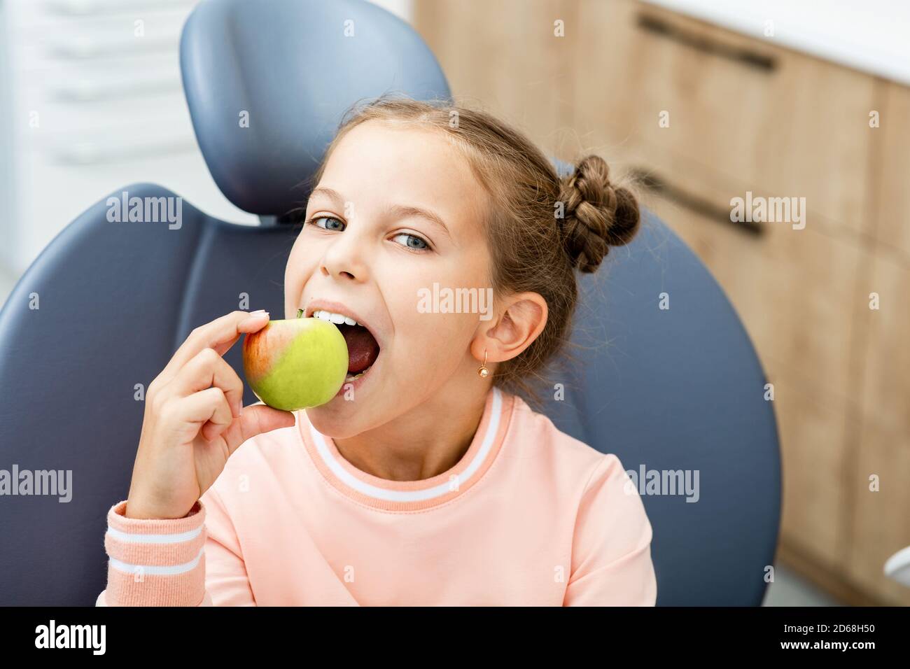Soins des dents pour enfants. Fille souriante piquant une pomme, dans la chaise du dentiste. Banque D'Images