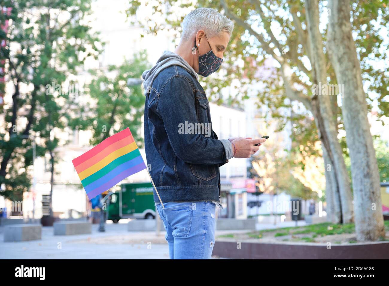 Jeune homosexuel caucasien portant un masque facial avec un drapeau LGBT dans sa poche discutant sur son smartphone. Célébration de la fierté LGBT en temps de pandémie. Banque D'Images
